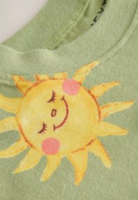 Camiseta de algodón verde claro con un gráfico de sol amarillo con una cara sonriente, mejillas rosas y rayos de sol ondulados. Textura suave.