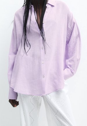 Personne portant une chemise ample lavande à boutons avec manches longues et un pantalon blanc orné de motifs en forme de cœur ajouré et de vagues.