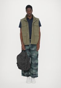 Gilet senza maniche verde con diverse tasche, camicia blu sotto, pantaloni mimetici e uno zaino nero decorato tenuto in una mano. Sneakers bianche.