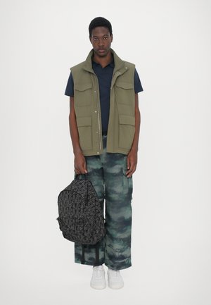 Gilet sans manches vert avec plusieurs poches, chemise marine en dessous, pantalon camouflage et un sac à dos noir à motifs tenu d'une main. Baskets blanches.
