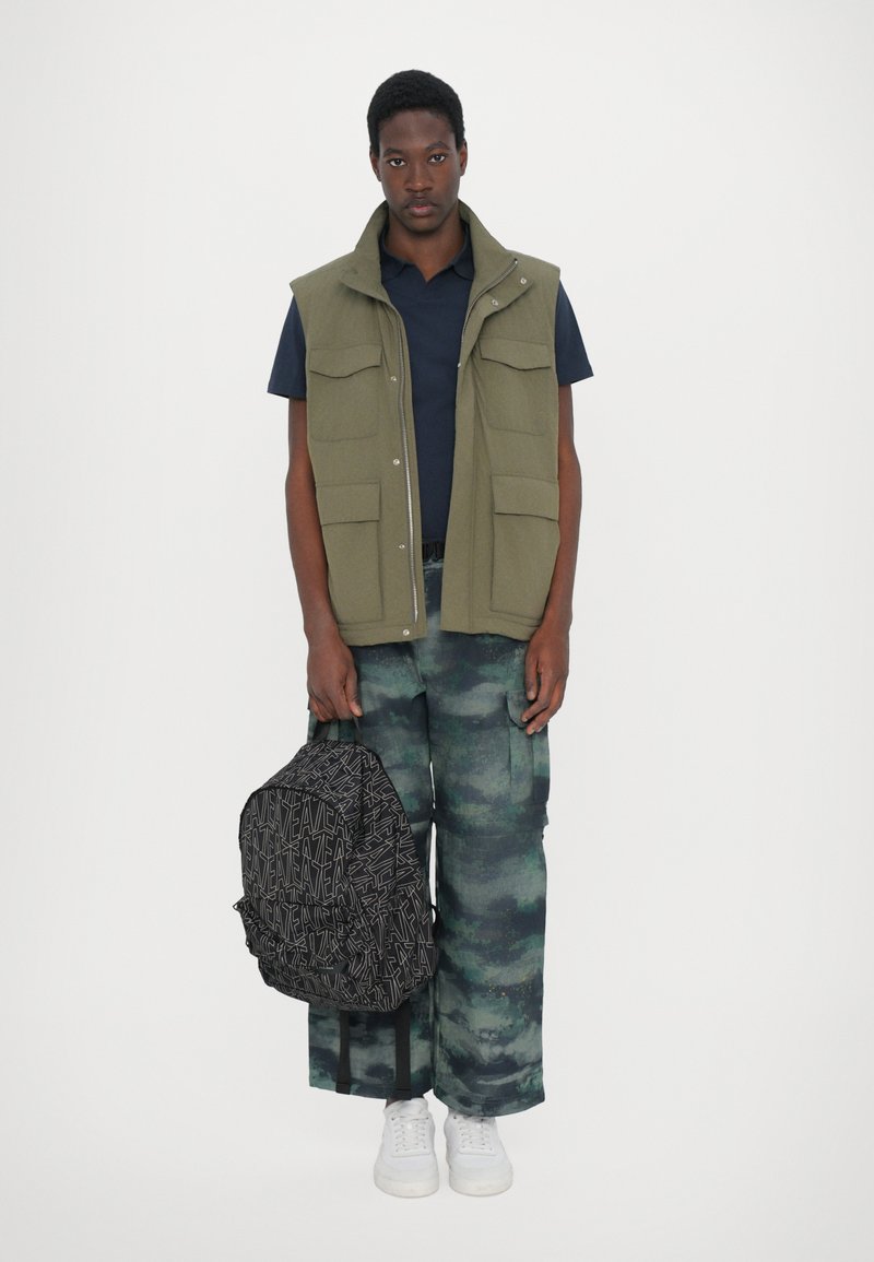 Gilet senza maniche verde con diverse tasche, camicia blu sotto, pantaloni mimetici e uno zaino nero decorato tenuto in una mano. Sneakers bianche.