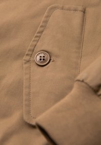 Tissu marron avec rabat de poche cousu et bouton rond couleur fauve embossé de « OMBRE » sur une manche de vêtement.