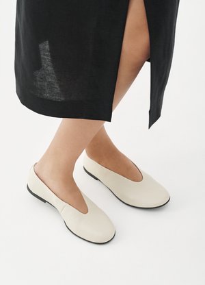 Osoba nosząca kremowe wsuwane buty z czarną podeszwą oraz czarną spódnicę z przednim rozcięciem, stojąca na białej powierzchni.