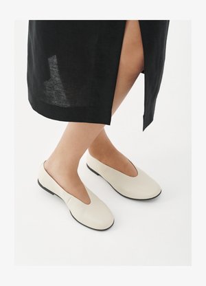 Άτομο που φοράει υποδήματα τύπου slip-on εκρού χρώματος με μαύρες σόλες και μαύρη φούστα με σχίσιμο μπροστά, στέκεται σε λευκή επιφάνεια.