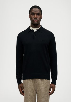 Pull polo en tricot noir avec col et trois boutons, arborant un logo discret. Porté par-dessus une chemise intérieure crème, associé à un pantalon texturé.