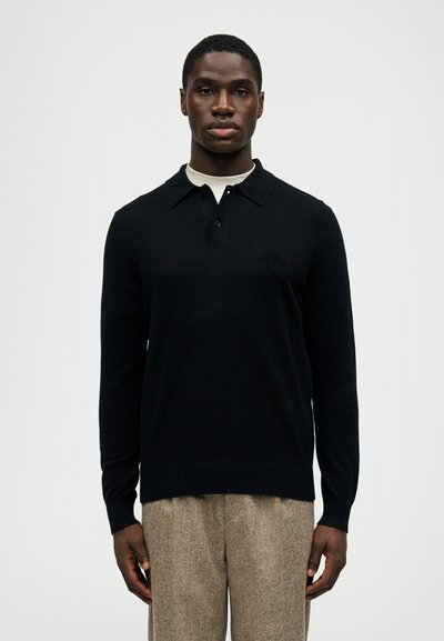 Schwarzer Strick-Polo-Pullover mit Kragen und drei Knöpfen, mit einem dezenten Logo. Über einem cremfarbenen Hemd getragen, kombiniert mit strukturierten Hosen.