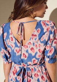 Fließendes Kleid in Blau mit pink-weißer abstrakter Musterung; verfügt über einen V-Ausschnitt, Bindeband und kurze Puffärmel. Glatte Stofftextur.