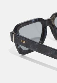 Schwarze marmorierte Sonnenbrille mit grauen Gläsern. Dickes, kantiges Gestell mit goldenen Akzenten und der Gravur "R.S.F" am Bügel. Glatte Textur.