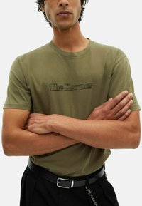 Hombre vestido con una camiseta verde oliva con el texto "The Kooples", pantalones negros, cinturón negro y cadena, con los brazos cruzados sobre el pecho.