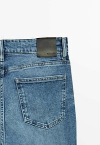 Vue arrière d'un jean en denim bleu montrant une seule poche arrière avec des coutures et une patch en cuir rectangulaire noir avec le logo de la marque sur la ceinture.