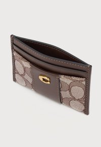 Λεπτή καφέ δερμάτινη θήκη για κάρτες με υφασμάτινες επιφάνειες με σχέδια, χρυσό κούμπωμα και πολλαπλές θήκες για κάρτες, γωνιακά τοποθετημένη σε λευκό φόντο.