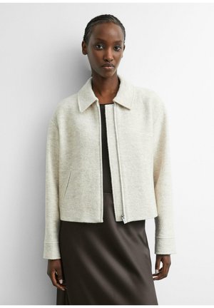 Woman with braided hair wearing a light beige zip-up jacket over a dark brown dress, standing against a plain white background.