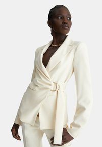 Blazer crema con vestibilità sartoriale, vita cintrata e revers affilati. Tessuto liscio, maniche lunghe e due tasche frontali.