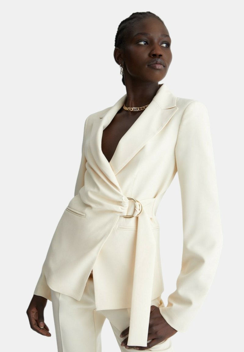 Blazer crema con vestibilità sartoriale, vita cintrata e revers affilati. Tessuto liscio, maniche lunghe e due tasche frontali.