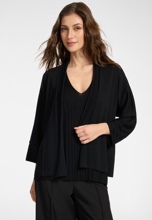 Kobieta o falujących brązowych włosach, ubrana w czarny plisowany top z dekoltem w serek oraz luźny czarny kardigan, pozująca na jednolitym jasnym tle.