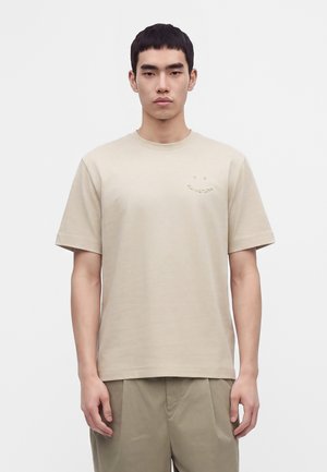 Homme asiatique portant un t-shirt beige uni à manches courtes et un pantalon marron clair, debout devant un fond blanc, face vers l'avant.