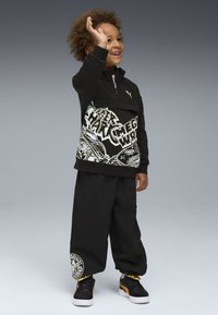 Bambino sorridente con i capelli ricci che saluta, indossando una felpa nera con grafica, pantaloni larghi neri e sneaker Puma nere con accenti arancioni.