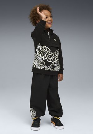 Lächelndes Kind mit lockigem Haar winkt, trägt einen schwarzen Sweatshirt mit Grafik, weite schwarze Hosen und schwarze Puma-Sneaker mit orangefarbenen Akzenten.
