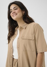 Chemise boutonnée beige clair à manches courtes, avec une poche poitrine, texture douce, portée sur un haut blanc. Design simple et coupe décontractée.