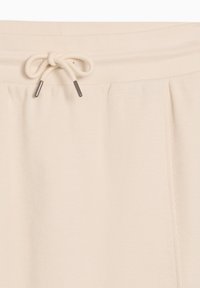 Gros plan sur la ceinture d'un pantalon en tissu beige texturé avec un cordon de serrage noué orné d'embouts métalliques.