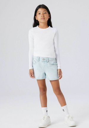 Ung pige står vendt fremad iført en hvid langærmet trøje, lyseblå denimshorts, hvide sokker og hvide sneakers på en ensfarvet baggrund.