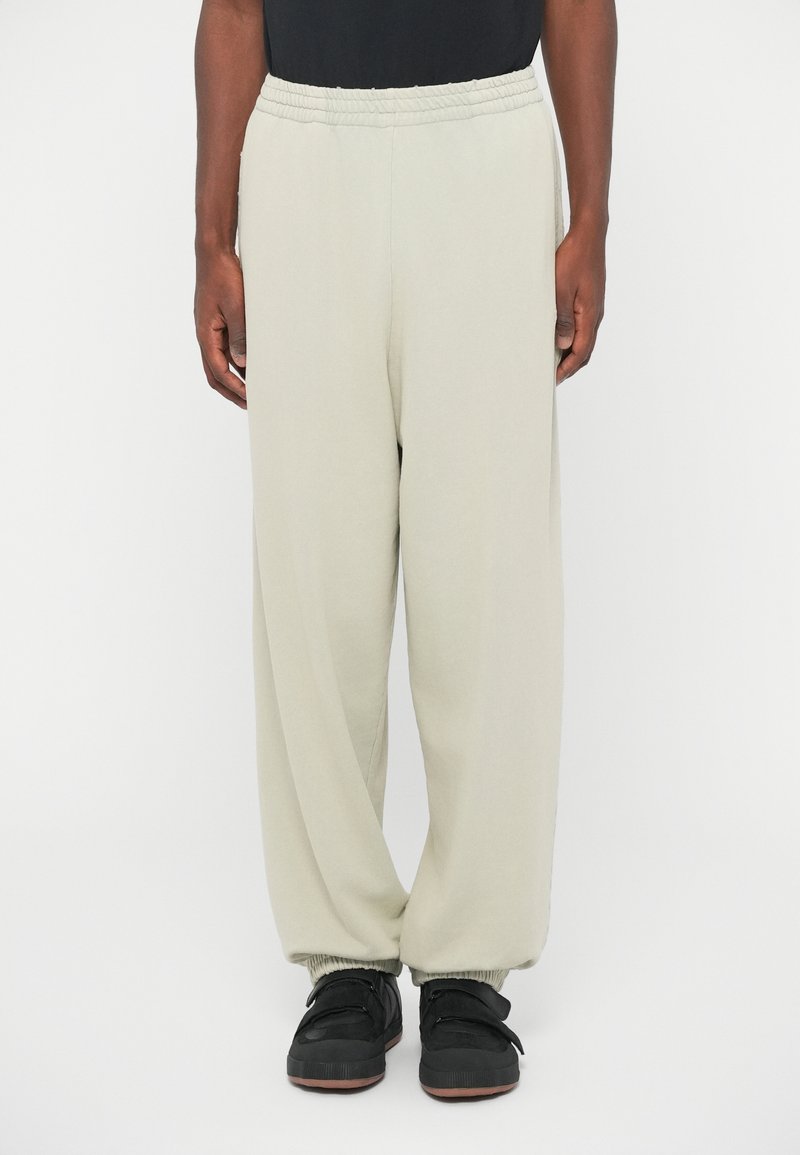Personne portant un pantalon de survêtement élastique beige clair avec des poignets froncés et des baskets noires sur un fond blanc uni.