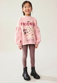 Felpa rosa con maniche voluminosi e testo grafico, abbinata a leggings marroni e stivaletti neri. Lo sfondo è neutro e uniforme.