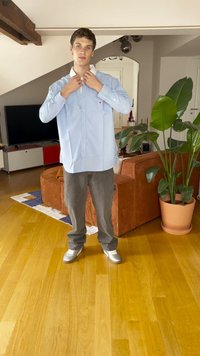 Jasnoniebieska koszula z długim rękawem i białym kołnierzykiem, szare spodnie i srebrne trampki. Stoi w pokoju z brązową sofą i roślinami.