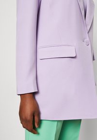 Blazer lavande à fermeture à un seul bouton, avec un revers cranté et une poche latérale, porté avec un pantalon vert menthe. Texture lisse.