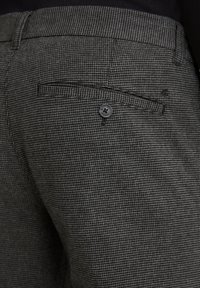 Nahaufnahme von grauen Hahnentrittmusterhosen mit einer rückseitigen, mit Knopf versehenen Paspeltasche und Gürtelschlaufen.
