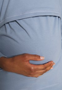 Indumento azzurro chiaro e elasticizzato, con un pannello arricciato sopra un ventre arrotondato, accompagnato da una mano slanciata appoggiata sul tessuto.