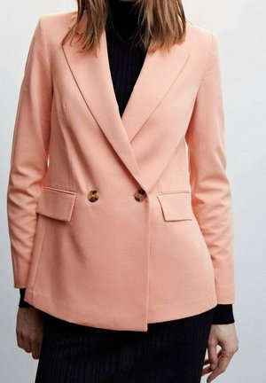 Woman wearing a light pink double-breasted blazer with tortoiseshell buttons over a black turtleneck and black skirt.