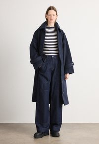 Marineblå ullkåpe med stor krage, stripete svart-hvitt langermet topp og vide denimbukser, komplettert med kraftige svarte sko.