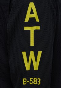 Schwarzer Stoff mit großen gelben Buchstaben "A", "T", "W" und der Zahl "B-583". Der Stoff hat ein strukturiertes, netzartiges Aussehen.