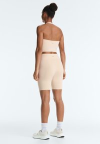 Femme avec les cheveux en chignon, portant un haut court beige à col halter et un short de cycliste assorti, baskets blanches, debout dos à la caméra.