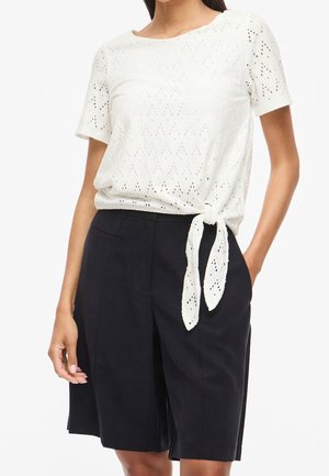 Femme portant un haut blanc à manches courtes en broderie anglaise, noué à la taille, avec un short noir taille haute arrivant aux genoux.
