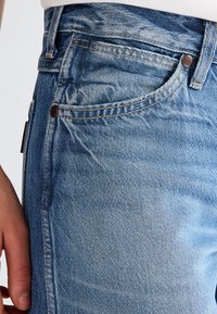 Close-up of blue denim jeans pocket with rivets, stitching details, and a person's left hand resting near the hip.