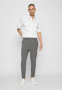 Jeune homme debout, mains dans le dos, portant une chemise blanche à manches courtes, un pantalon gris à fines rayures avec chaîne, et des baskets blanches sur fond uni.