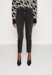 Person som bär högmidjade svarta skinny jeans med ett smalt svart bälte och svarta spetsiga klackskor, kombinerat med en svartvit mönstrad tröja.