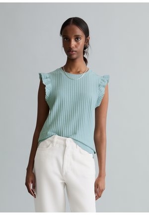 Kobieta ubrana w jasnoniebieski prążkowany top bez rękawów z falbankami na ramionach oraz białe, wysokie spodnie, stojąca na tle szarego tła.