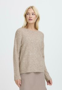 Jasno beżowy sweter z miękkiego, teksturowanego materiału, z okrągłym dekoltem, długimi rękawami i prążkowanymi mankietami, noszony z plisowaną spódnicą.