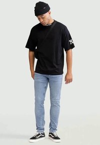 T-shirt oversized preta com uma alça diagonal, jeans skinny azuis claros e ténis preto e branco. Um gorro preto completa o visual.