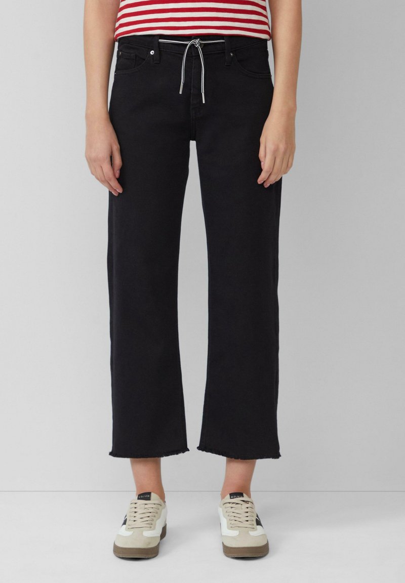 Personne portant un jean noir court à jambes larges avec des ourlets effilochés, un t-shirt rayé rouge et blanc, et des baskets blanches avec des semelles beiges.