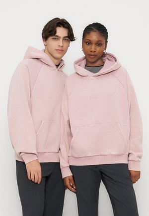 Δύο μοντέλα που φορούν τα ίδια ανοιχτό ροζ φούτερ με κουκούλα και μπροστινές τσέπες, καθώς και σκούρα παντελόνια, στέκονται κοντά σε λευκό φόντο.