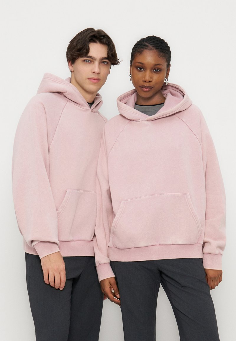 Dva modela, oblečena v ujemajoče se svetloroza potegnjene puloverje s kapuco in sprednjimi žepi ter temne hlače, stojita skupaj pred belim ozadjem.