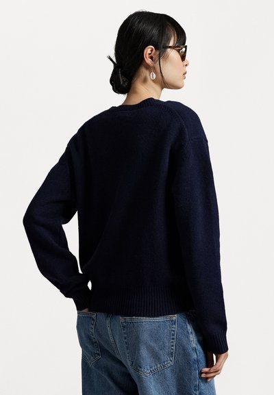 Navyblauwe oversized gebreide trui met geribde zoom en mouwen. Model draagt een lichtblauwe jeans en wordt van achteren getoond met een neutrale achtergrond.