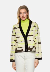 Gekürzter Cardigan in Creme, Braun und Neon-Grün Camouflage-Muster; mit einem schwarzen V-Ausschnitt und fünf Knöpfen auf der Vorderseite.