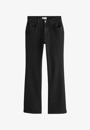 Osoba oblečená v čiernych džínsoch s vysokým pásom a bielom tričku s dlhým rukávom s čiernymi vertikálnymi pásikmi, ruka v zadnom vrecku.