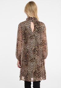 Frau mit blonden Haaren, die ein langärmliges Kleid mit Leopardenmuster und einem gerafften hohen Kragen sowie einem Schlüsselloch-Detail auf dem Rücken trägt, kombiniert mit schwarzen Hosen.