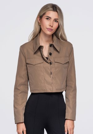 Jeune femme aux longs cheveux blonds portant une veste courte marron boutonnée et un pantalon noir taille haute sur un fond clair uni.